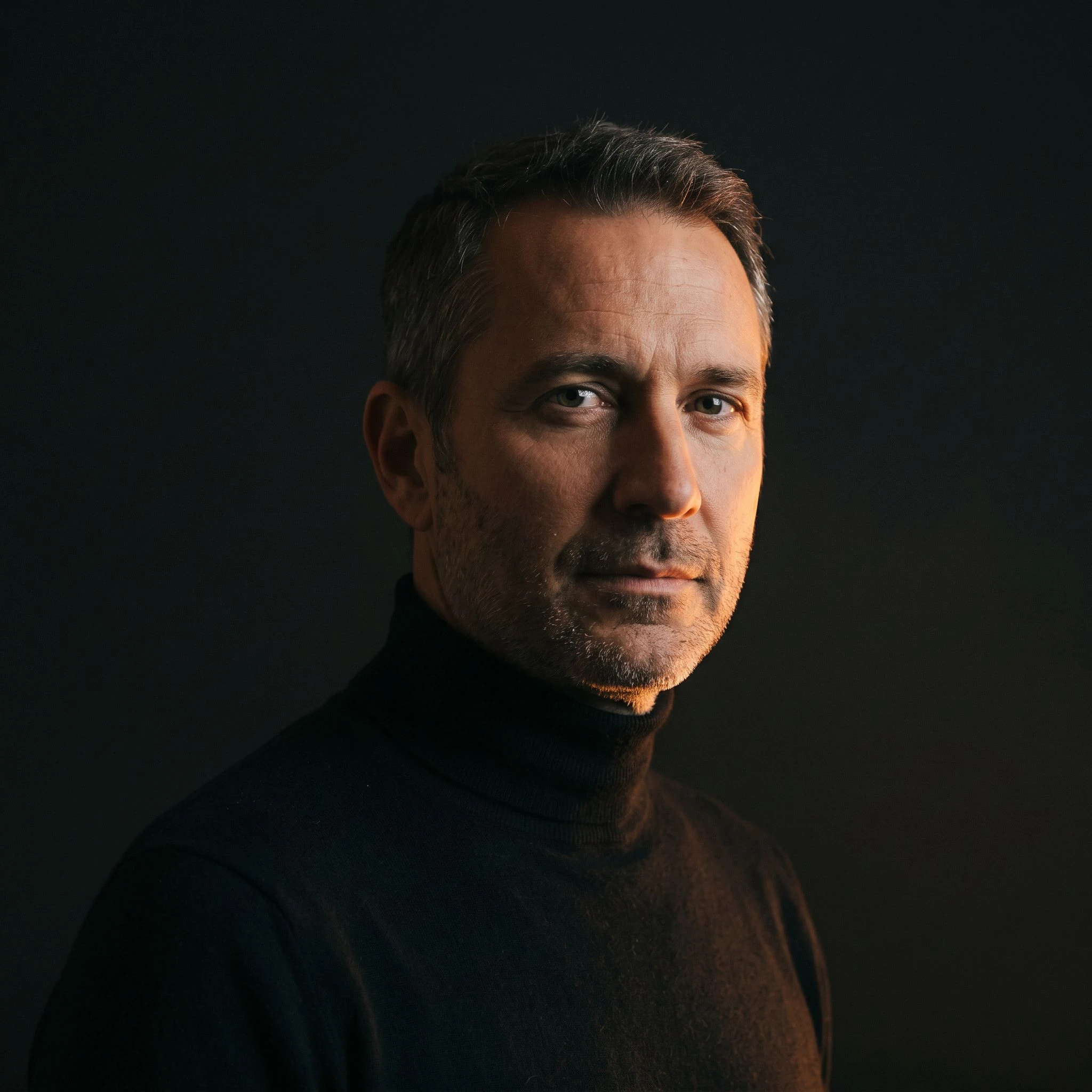 A candid editorial portrait of a professional man in his early fifties, lit from below-right with warm amber tones, against a near-black background